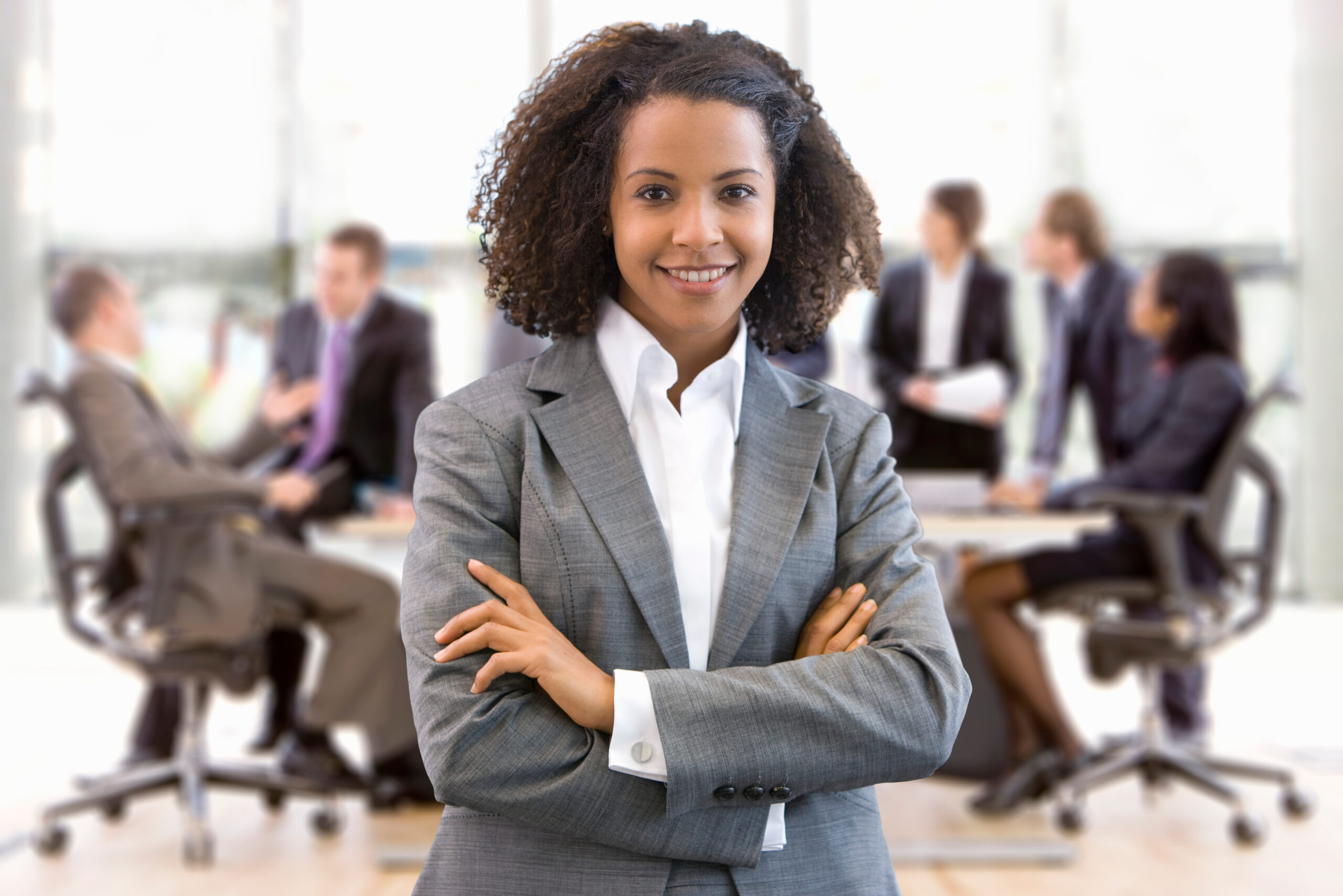 A young business professional standing confidently in a boardroom setting with her peers in the background.