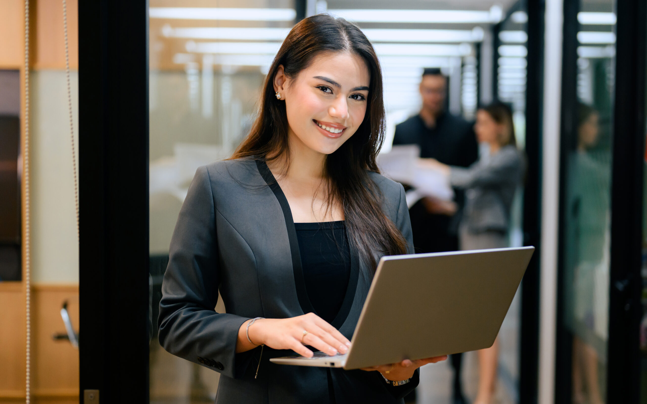 A young professional standing in a modern office setting while holding a laptop